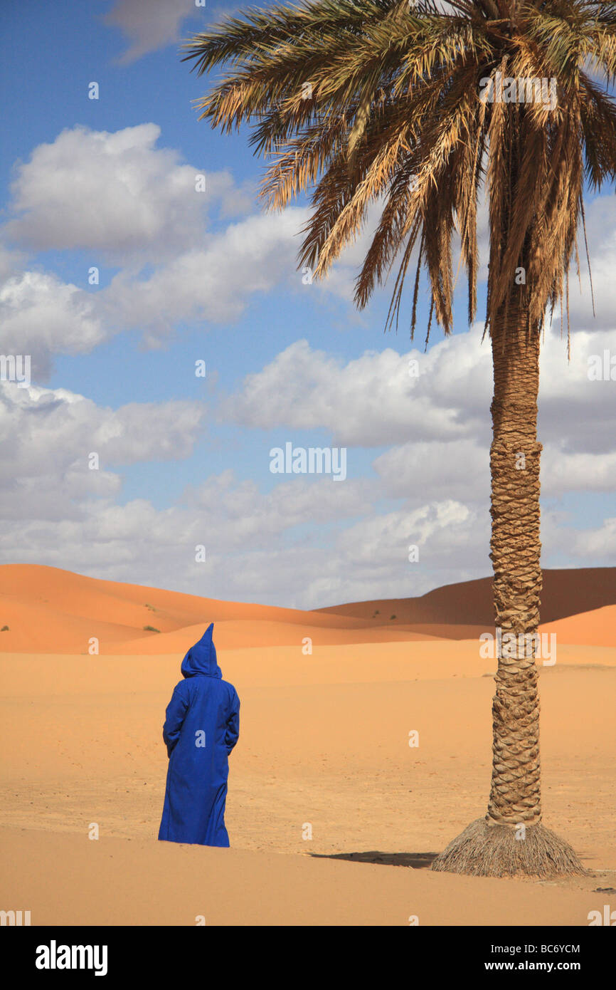 Persona che indossa Marocchino tradizionale djellabah sotto una palma in Erg Chebbi dune di sabbia del deserto del Sahara, Merzouga, Moroccco Foto Stock