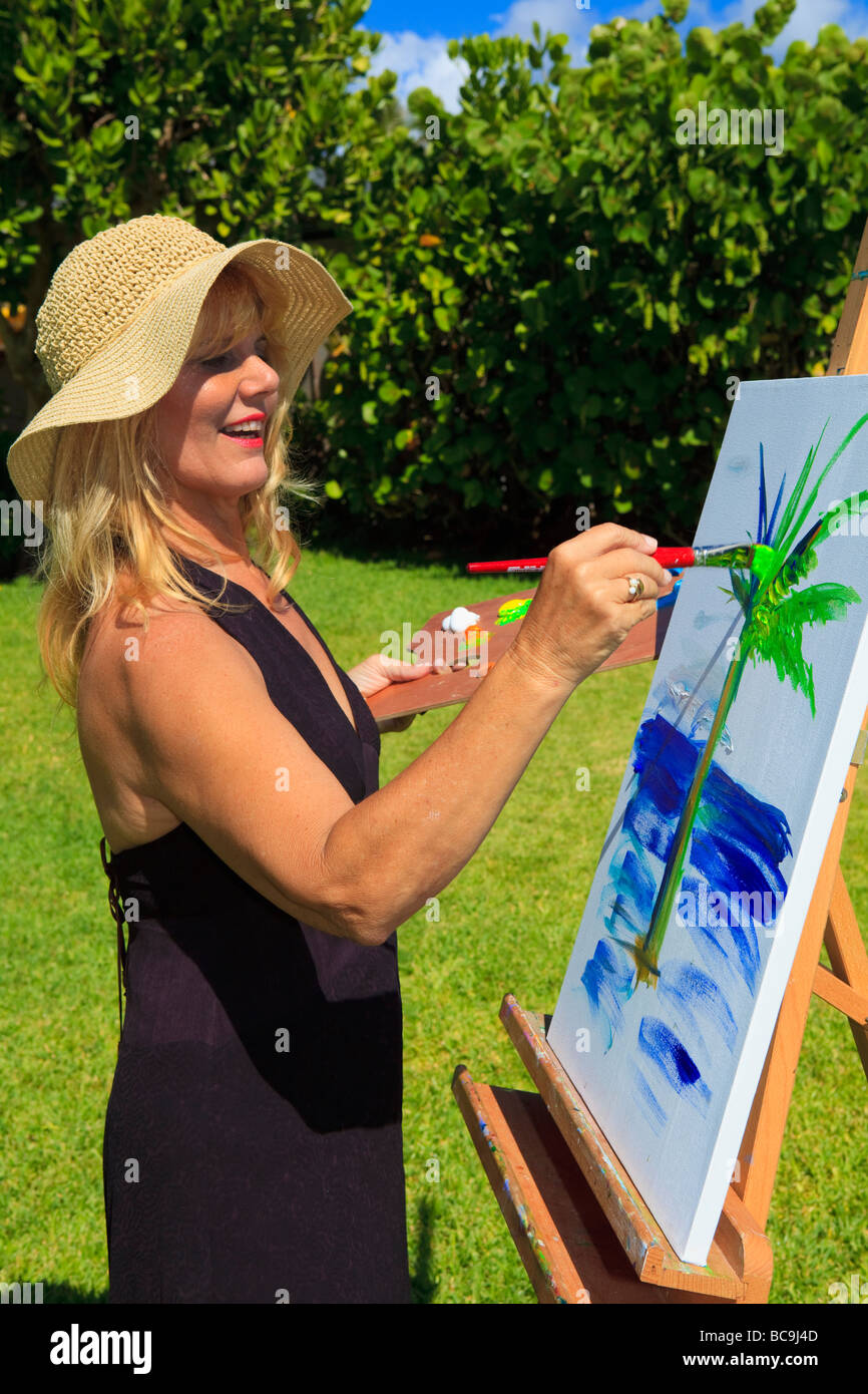 Un artista femminile nei suoi anni cinquanta creando un dipinto al di fuori su tela di una palma e l'oceano in Lanikai, Hawaii Foto Stock