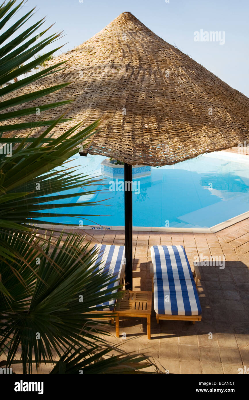 Guardando attraverso foglie di palmo di una coppia del legno e di lettini per il sole sotto un sole di vimini ombra dalla piscina Foto Stock
