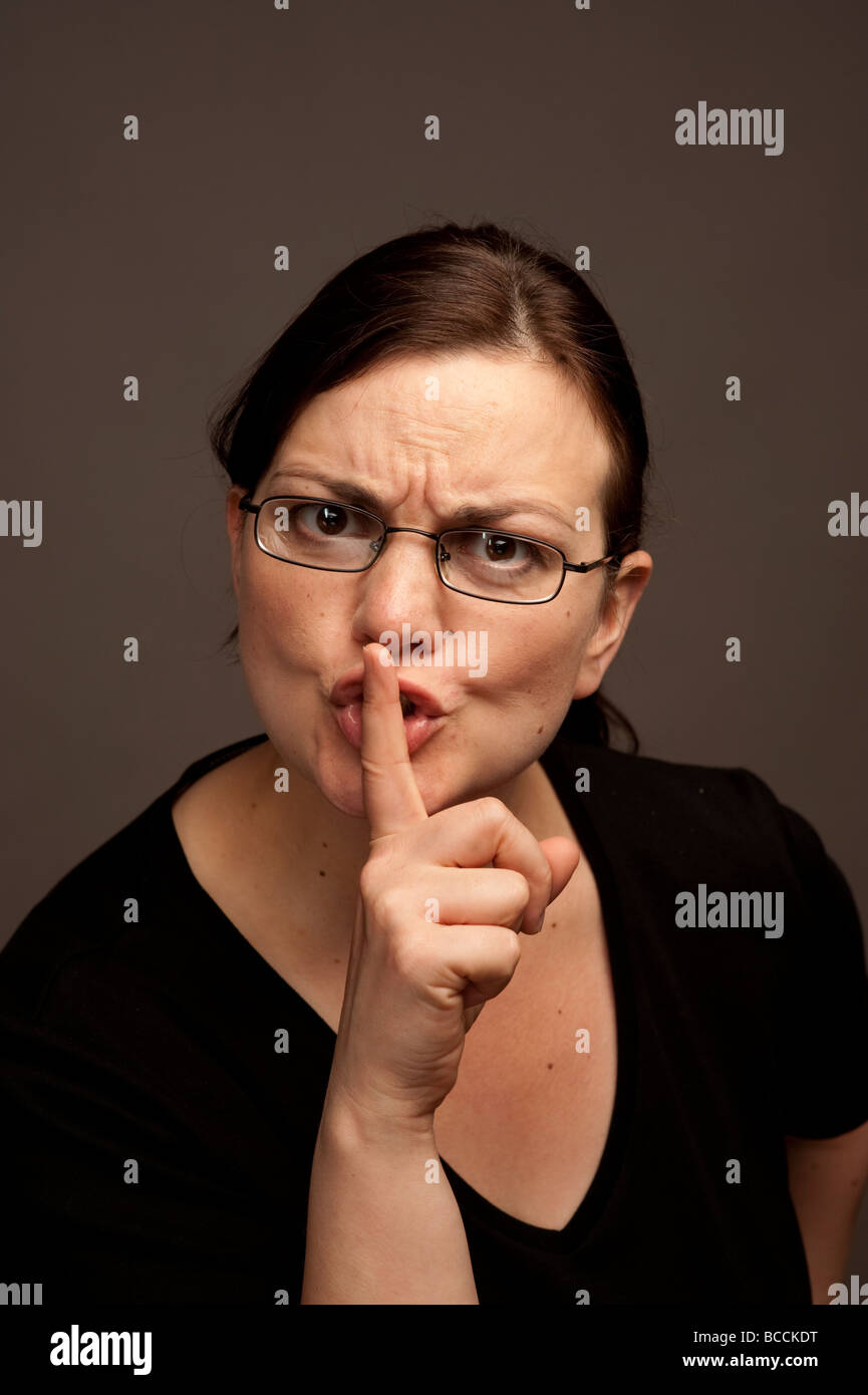 Una donna che indossa gli occhiali con il dito sulle labbra per chiedere silenzio non tranquilla parlando Foto Stock