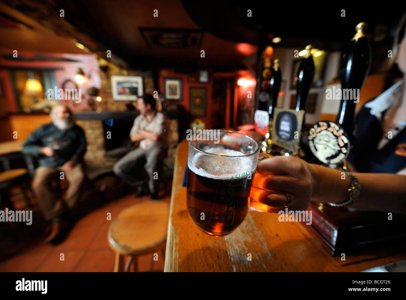 Due uomini seduti a un fuoco aperto in un pub tradizionale come la padrona di casa mette una pinta di birra al bar GLOUCESTERSHIRE England Regno Unito Foto Stock