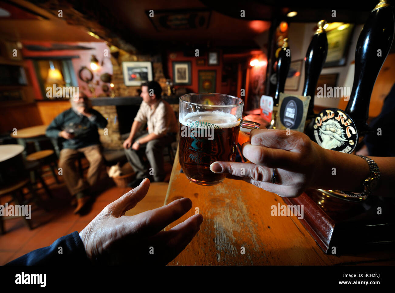 Due uomini seduti a un fuoco aperto in un pub tradizionale come la padrona di casa serve una pinta di birra al di sopra del bar GLOUCESTERSHIRE England Regno Unito Foto Stock