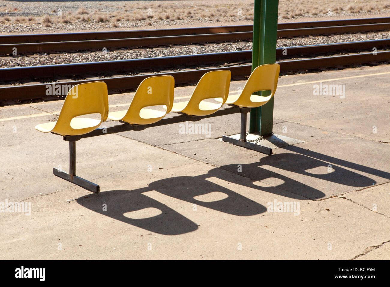 Fila di quattro sedie di colore giallo che gettano ombre sulla piattaforma del treno alla stazione ferroviaria con binari in background Foto Stock