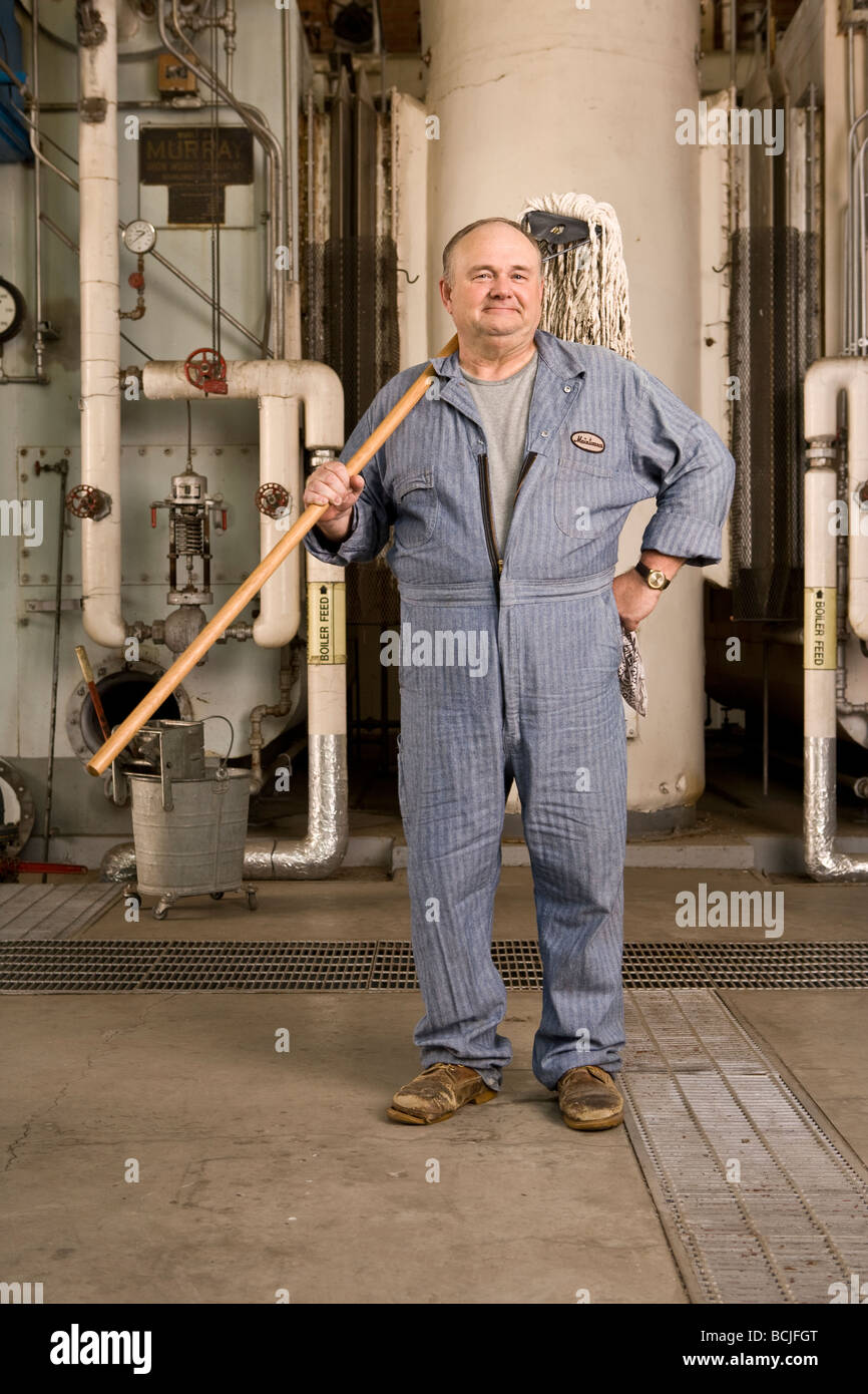 Caucasico maschio in blu custode/bidello tute in impostazione di fabbrica compresi caldaia, tubazioni, valvole. Egli è in possesso di mop. Foto Stock