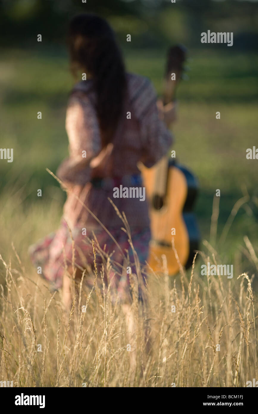 Vista posteriore della donna che cammina attraverso il campo, portando la chitarra, concentrarsi su erba alta in primo piano Foto Stock