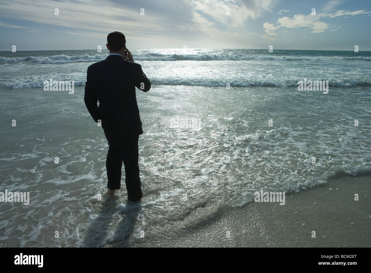 Imprenditore in piedi di surf in spiaggia, tramite telefono cellulare, vista posteriore Foto Stock