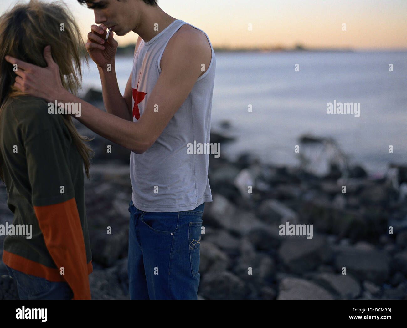 Coppia giovane in piedi faccia a faccia, fumare vicino a bordo di acqua, vista laterale Foto Stock