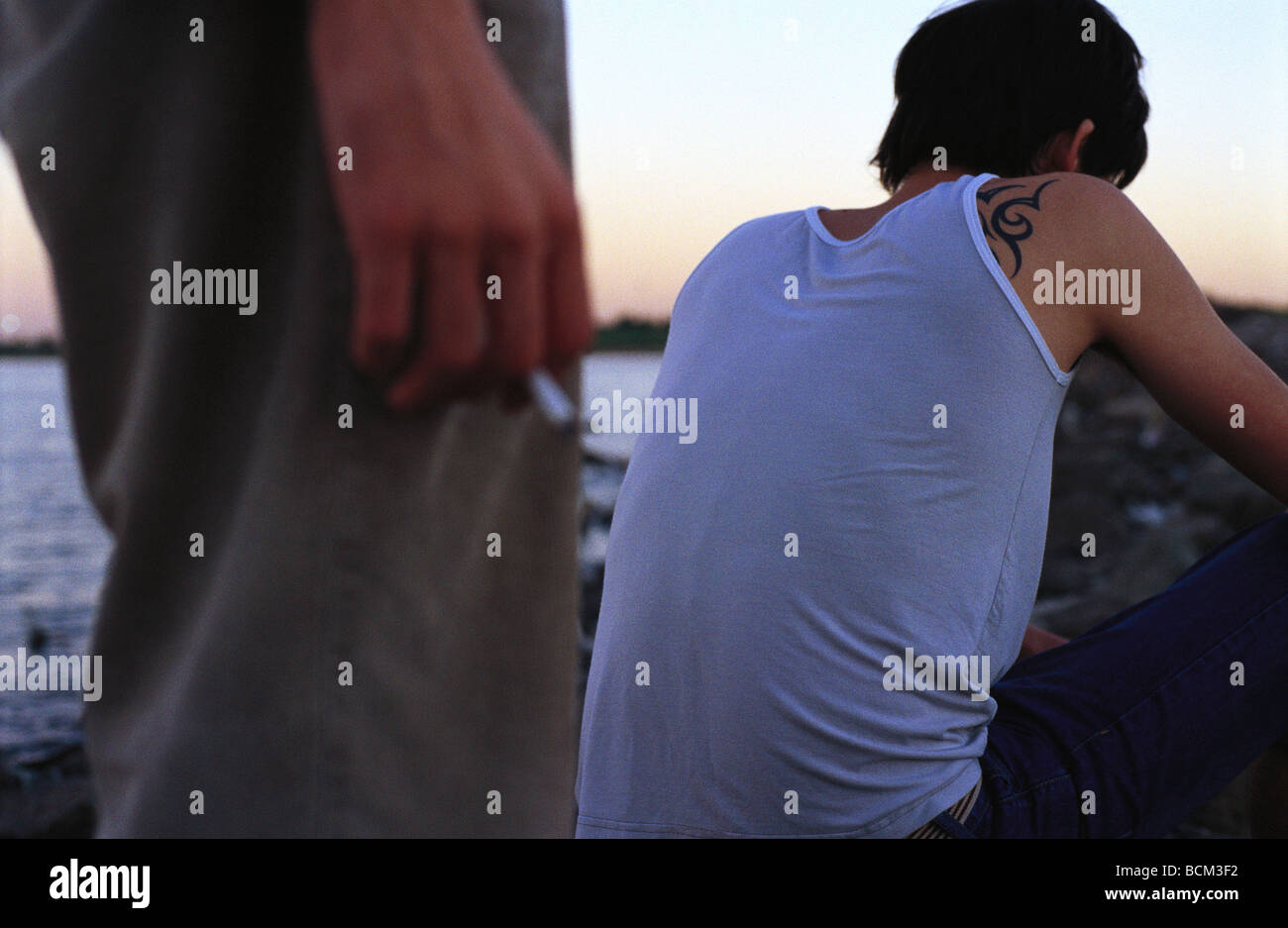 Due giovani uomini sulla riva, uno in piedi con la sigaretta, altri accovacciato, vista posteriore, ritagliato Foto Stock