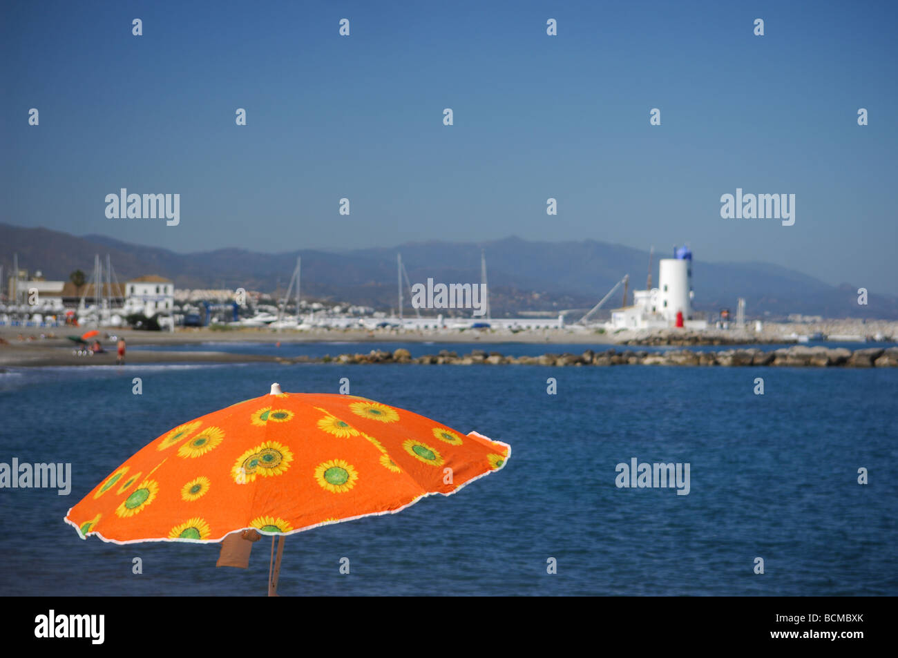 Un colore arancio ombrellone in primo piano cercando su un litorale spagnolo con un faro e yacht. Foto Stock