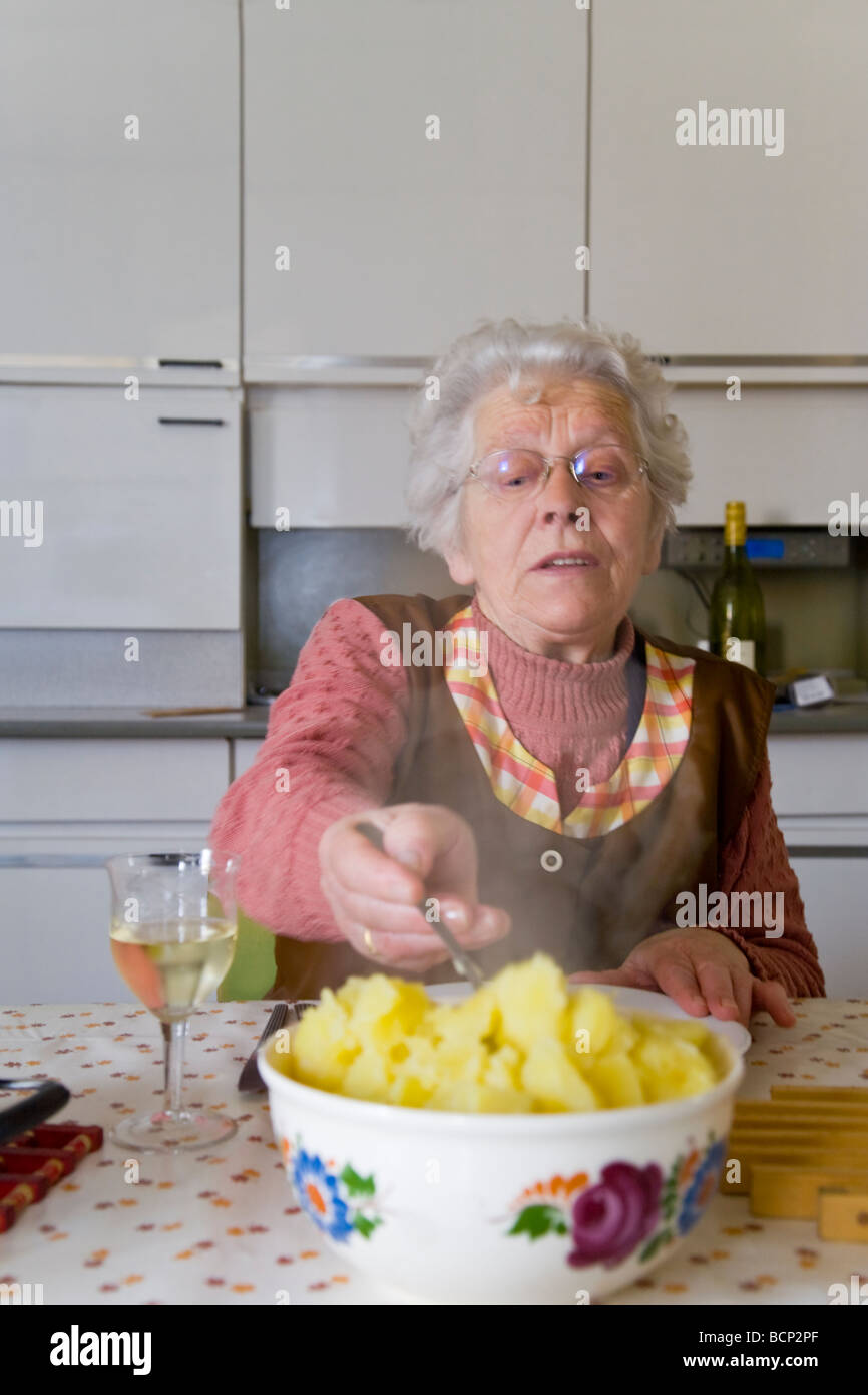 Frau in ihren Siebzigern sitzt in der Küche am gedeckten Mittagstisch und nimmt sich Kartoffeln auf Ihren Teller Foto Stock