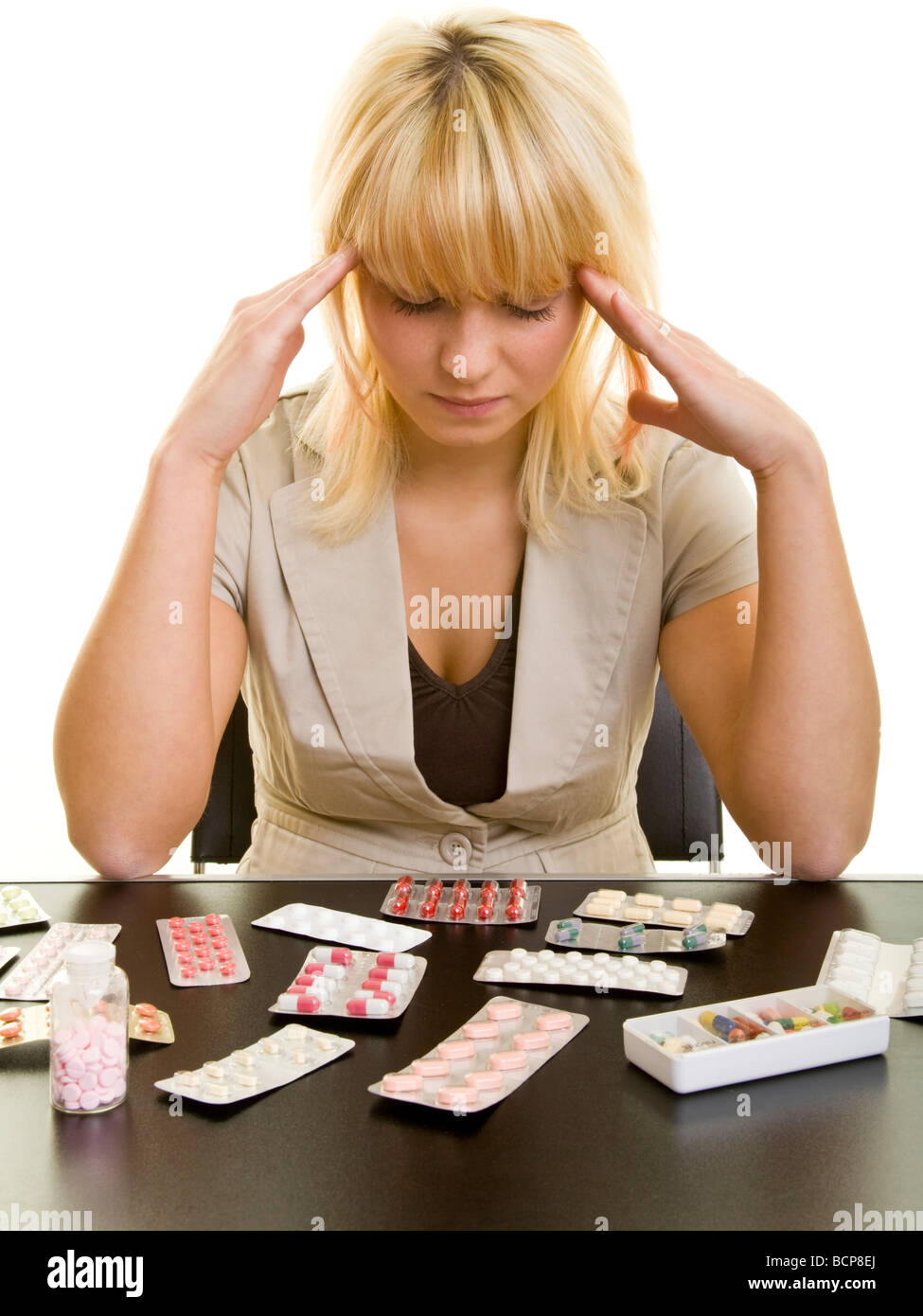 Junge Frau sitzt mit Kopfschmerzen vor einem Tisch mit vielen Arzneimitteln Foto Stock