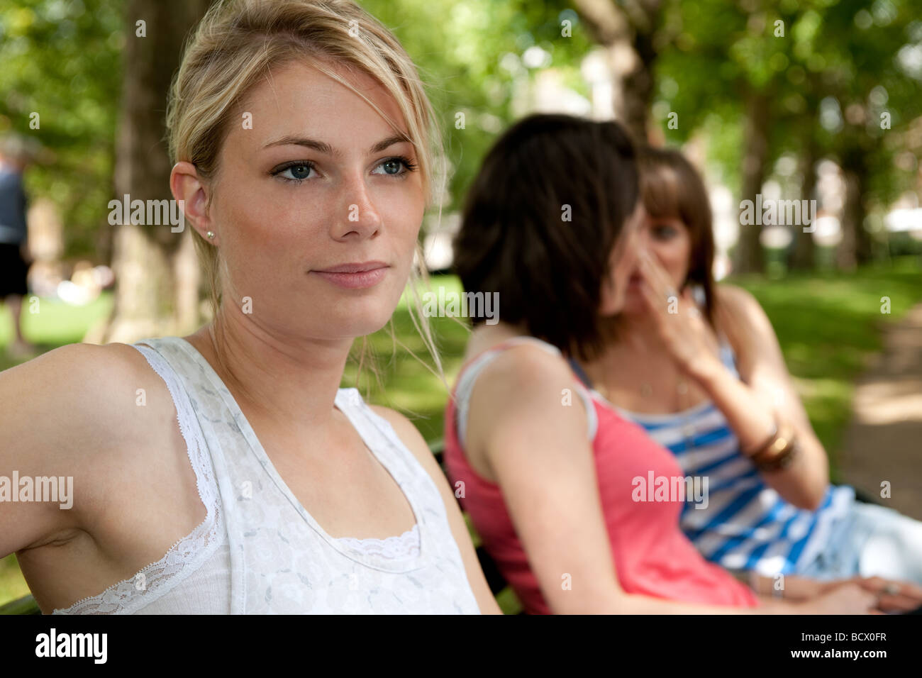 Due giovani ragazze sussurro a ogni altro circa una terza ragazza, tutti seduti su una panchina nel parco verde, Londra Foto Stock