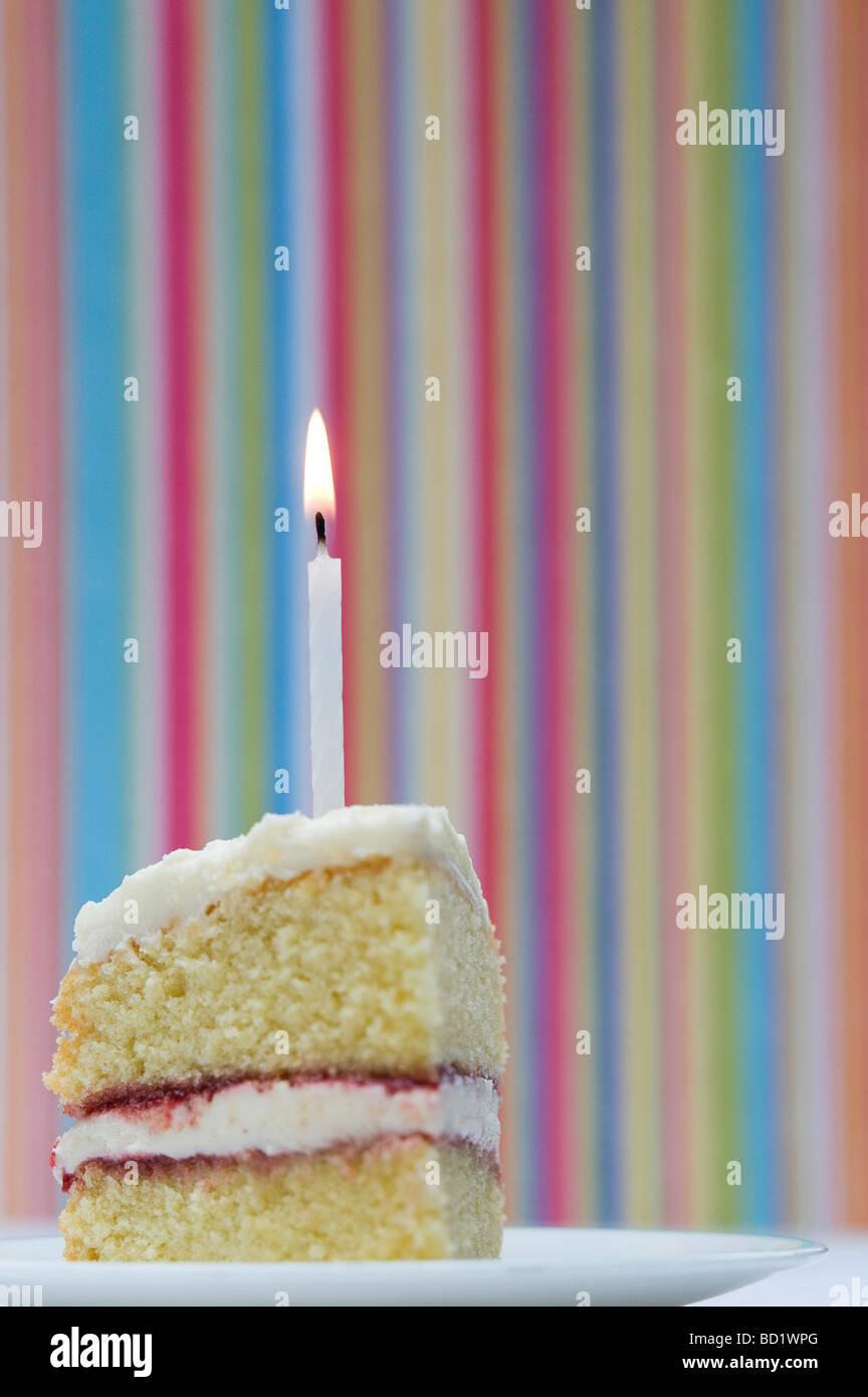 Fetta di torta di compleanno con una candela accesa contro un pastello colorato sfondo a strisce Foto Stock