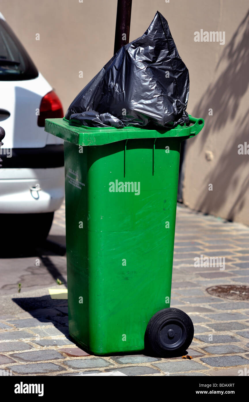 Green Wheely Bin si trova in strada, con un sacco di plastica traboccante / 'Garbage CAN' in strada, rifiuti di parigi, contenitori per rifiuti parigi Foto Stock