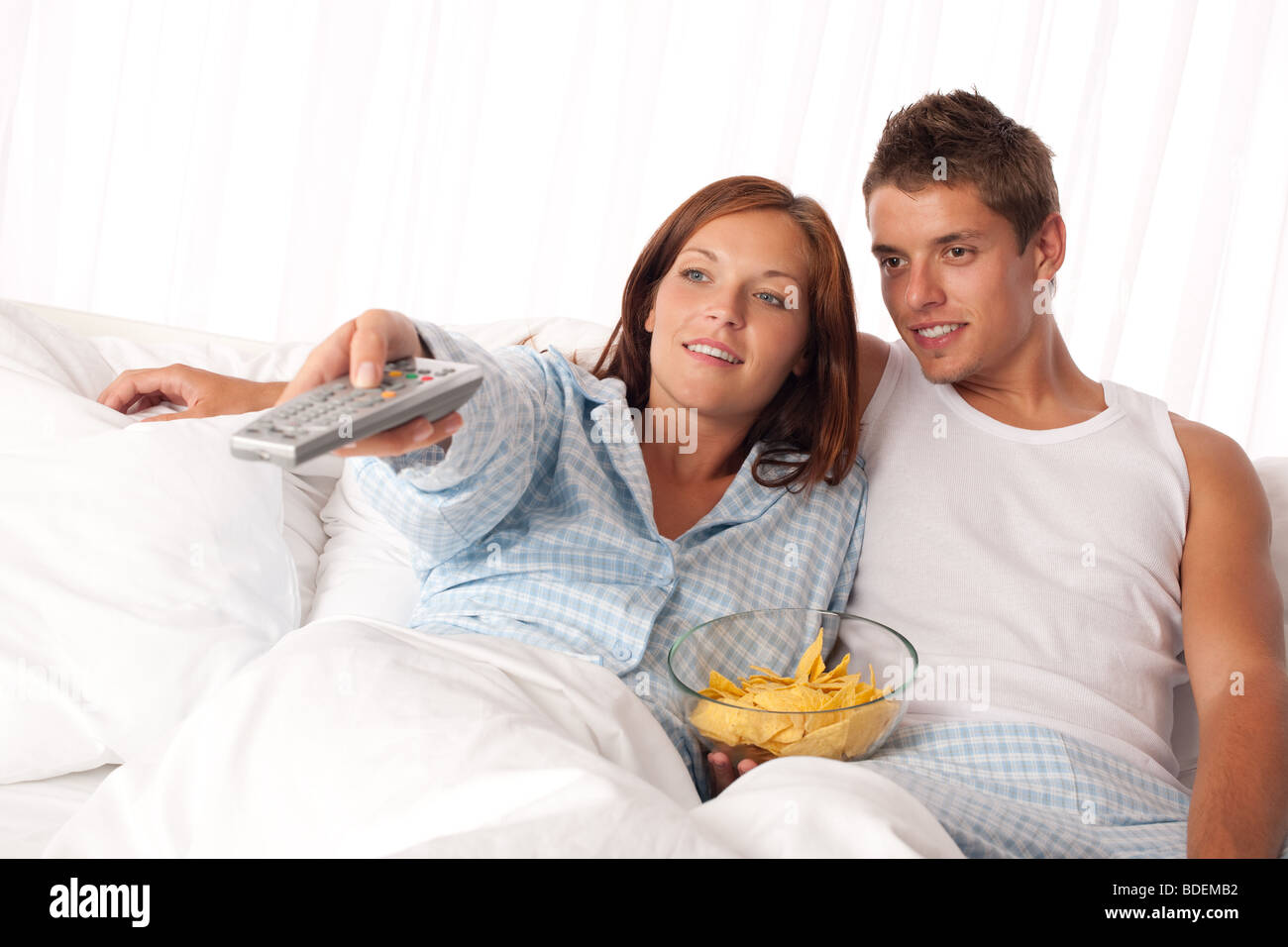 Giovane donna e uomo disteso nel letto a guardare la televisione e mangiare patatine Foto Stock