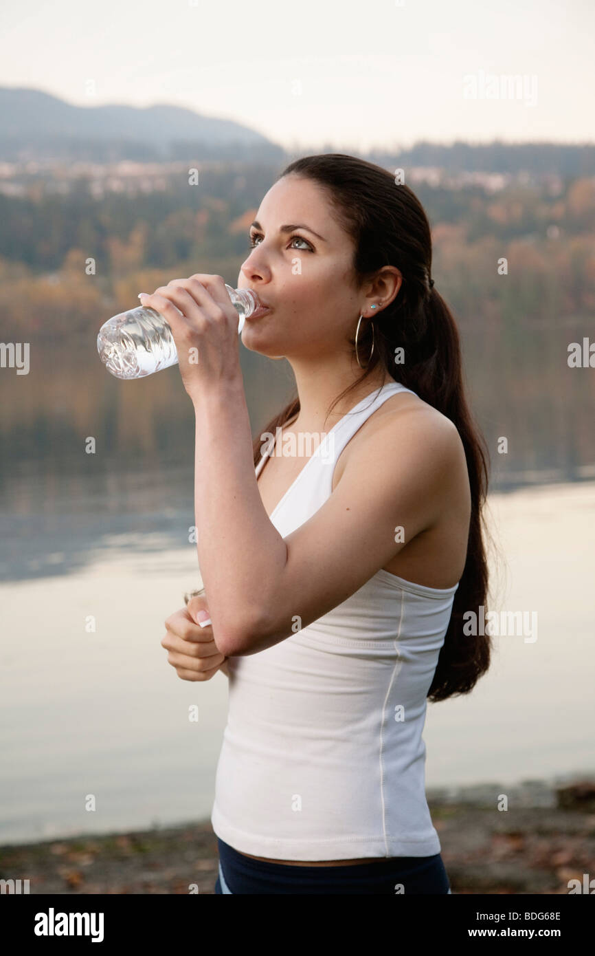 Una giovane donna atletica bere da una bottiglia d'acqua in plastica accanto a un lago. Foto Stock