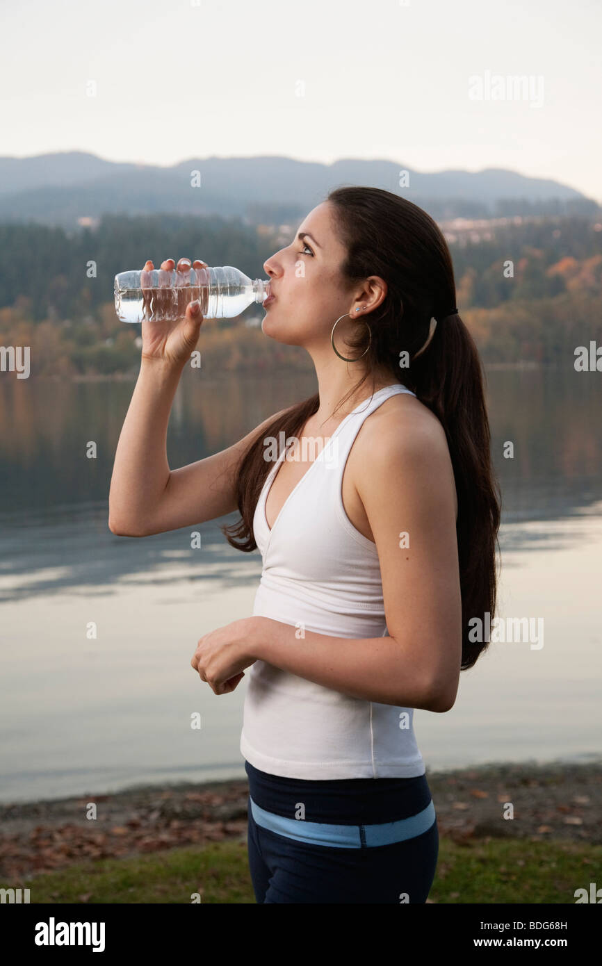 Una giovane donna atletica bere da una bottiglia d'acqua in plastica accanto a un lago. Foto Stock