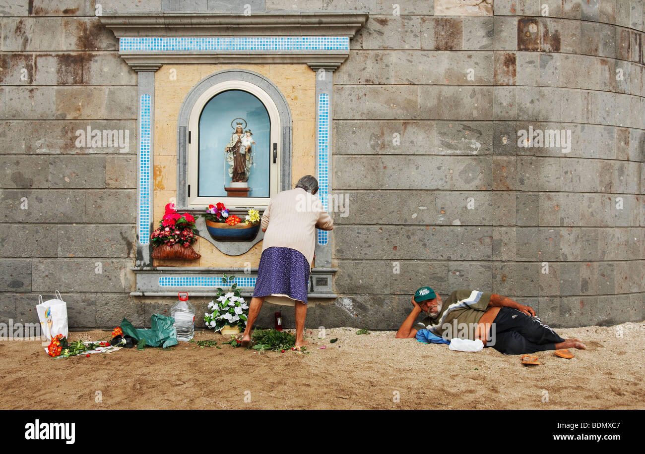 Il vecchio donna spagnola cambiando i fiori al santuario sulla spiaggia mentre l uomo orologi. Potrebbe essere utilizzato per il divario di retribuzione tra donne e uomini La parità retributiva... Foto Stock