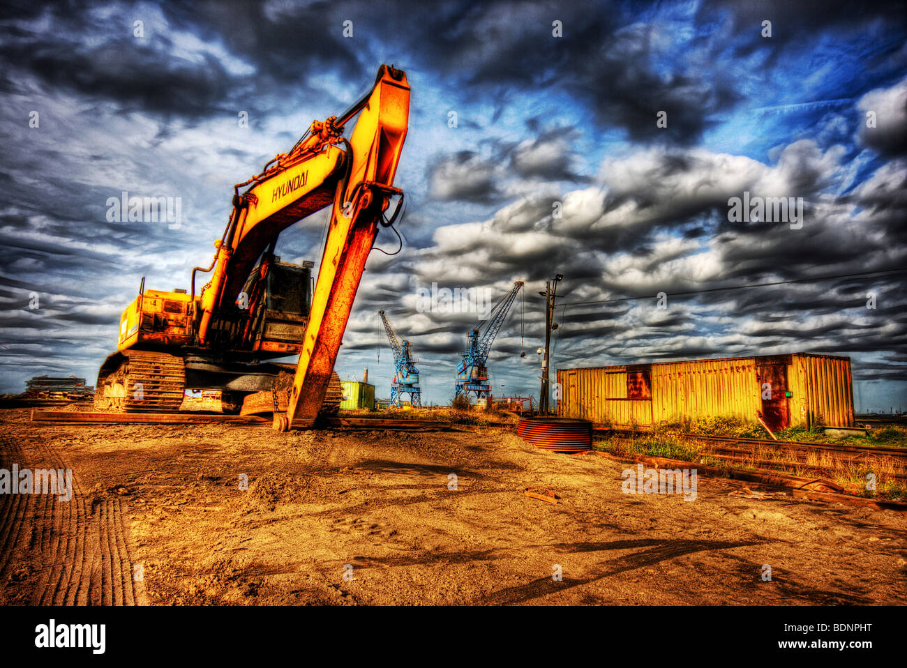 Grande giallo escavatore con cingoli sul sito di costruzione di gru in distanza e inquietanti nubi scure sotto un cielo blu Foto Stock