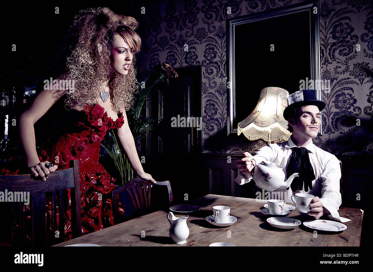 Una giovane donna che indossa un abito rosso guardando un giovane uomo seduto a un tavolo versando una tazza di tè che indossa un cappello a cilindro. Foto Stock