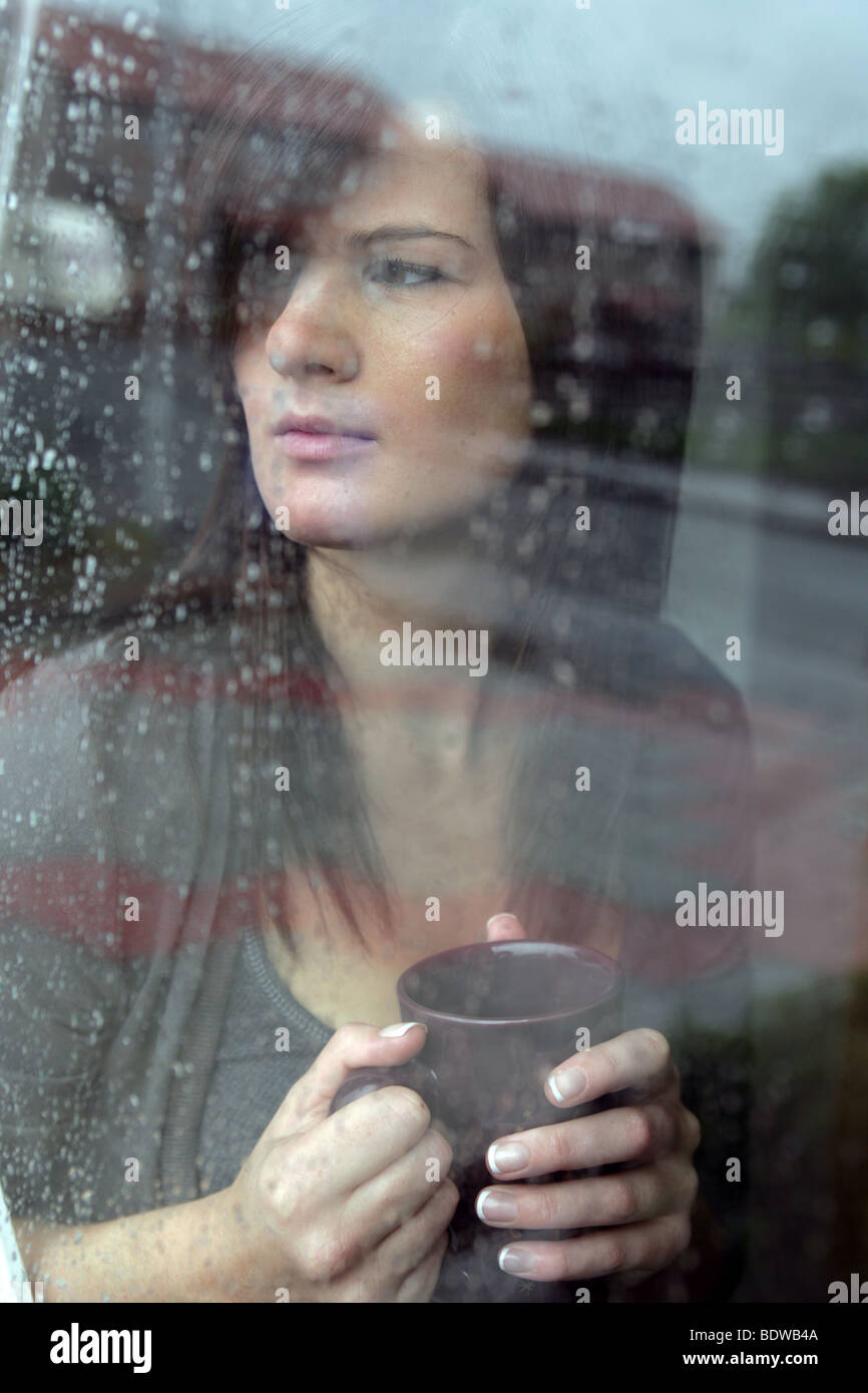 Dai capelli scuri donna tenendo una tazza di caffè guardando fuori una pioggia inzuppato finestra. Foto Stock