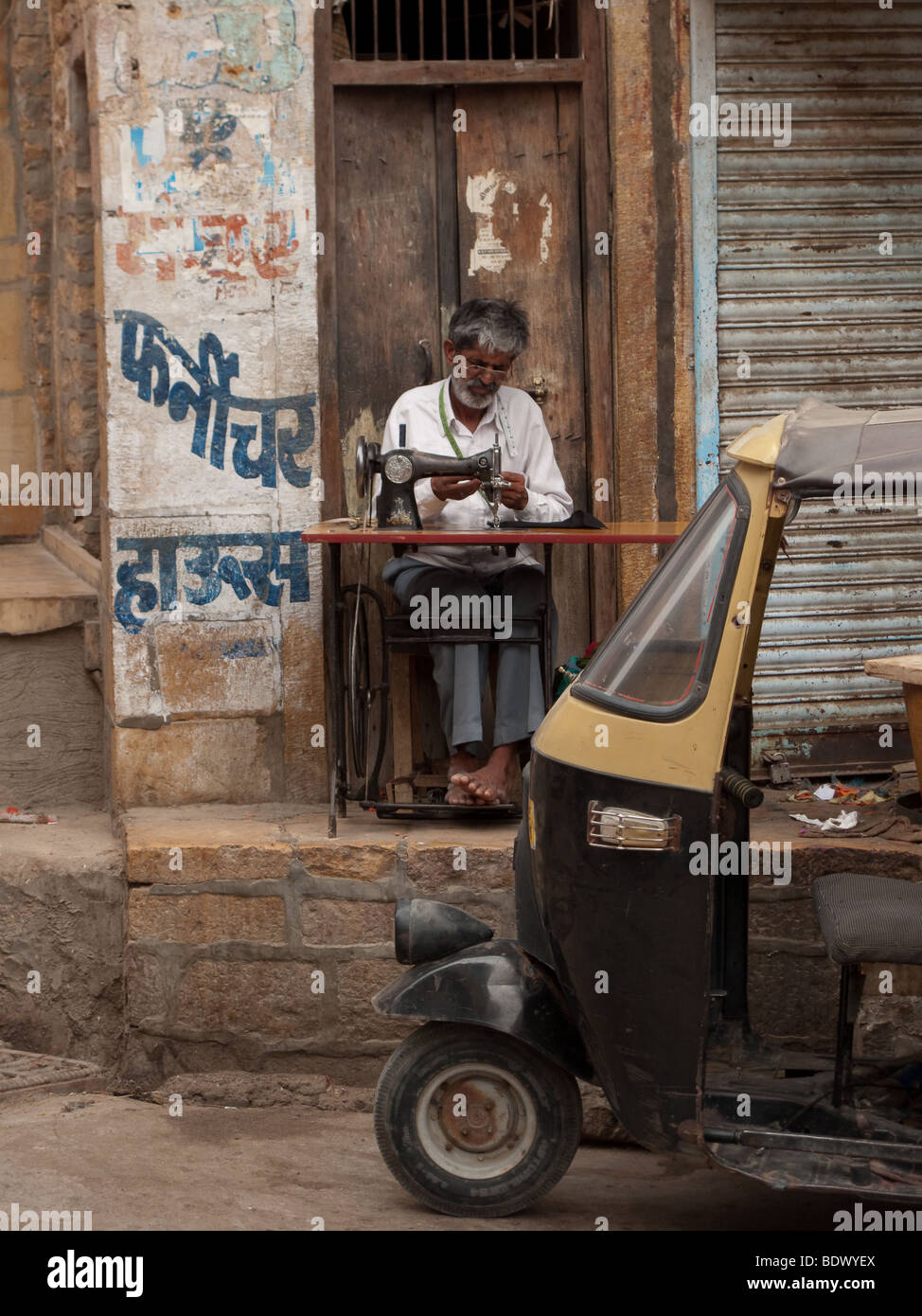 Jaisalmer, India - Un vecchio sarto imposta il suo negozio in un piccolo angolo sulla strada vicino a un rickshaw. Foto Stock