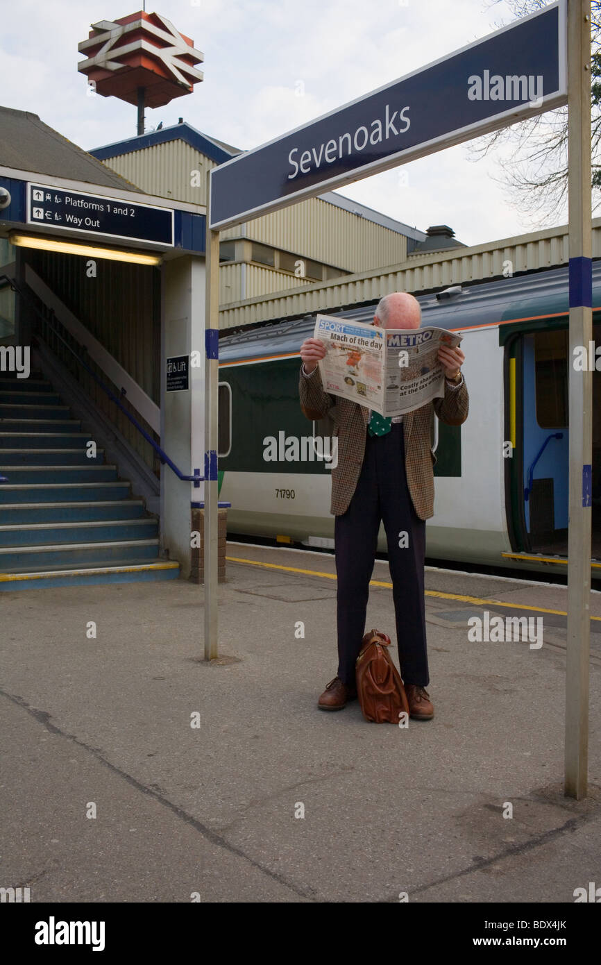 Un elegantemente vestito uomo anziano legge un giornale in attesa di un treno su un binario in stazione. Foto Stock