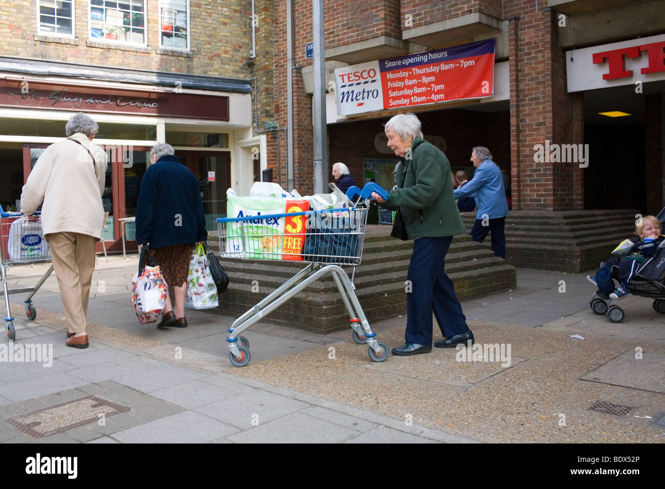 Una vecchia donna spinge un carrello pieno di shopping dal supermercato Tesco compreso un value pack di rotolo di carta igienica. Foto Stock