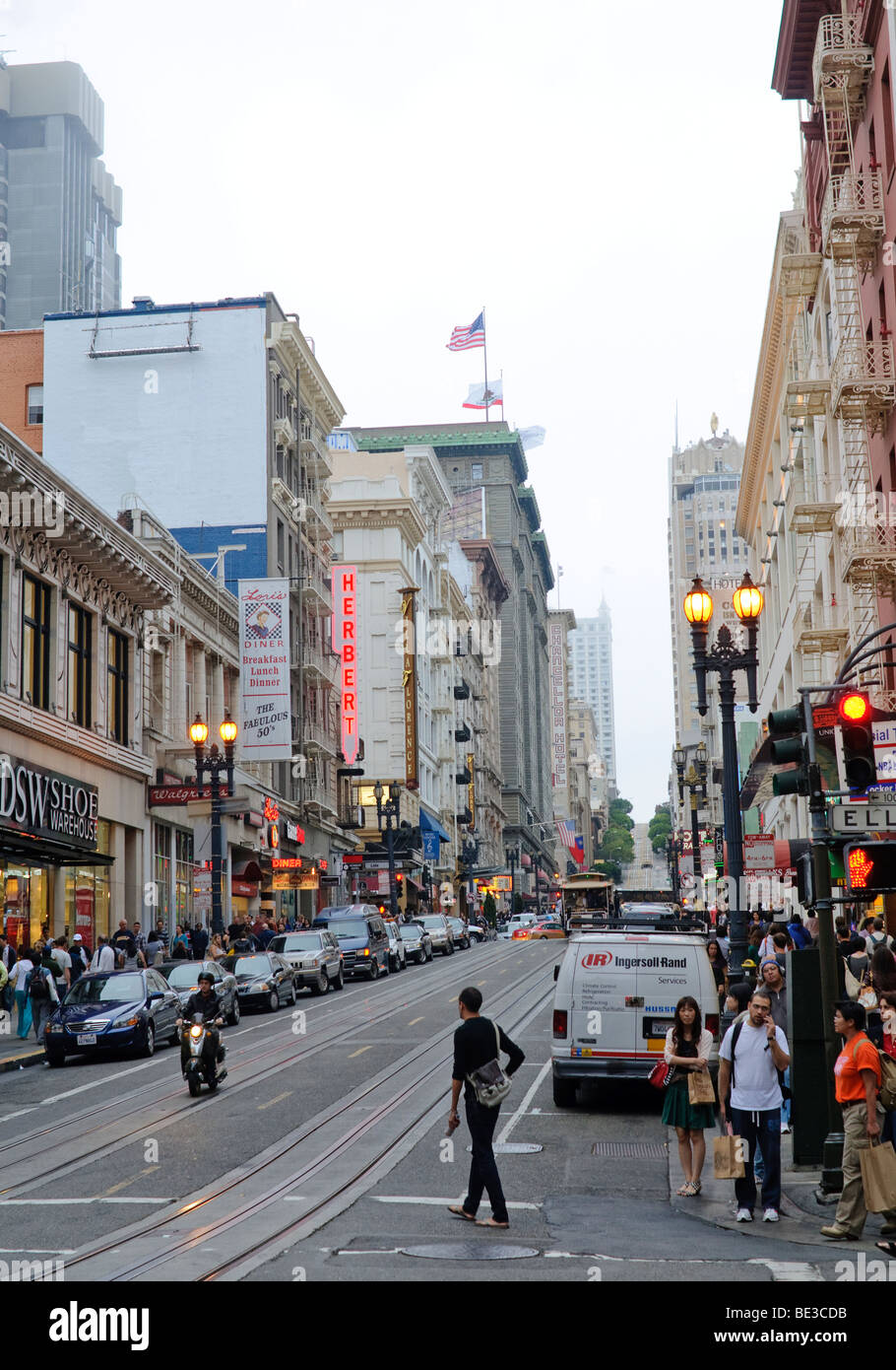 Piste per la funivia di Powell Street l'Herbert Hotel Chancellor Hotel San Francisco California // SAN FRANCISCO, California - Powell Street a San Francisco presenta iconici binari della funivia che corrono lungo il suo centro. La vivace strada è costeggiata da edifici storici, negozi e hotel di spicco, tra cui l'Herbert Hotel e il Chancellor Hotel. Powell Street è una parte vitale della funivia di San Francisco, un monumento storico nazionale e l'ultima rete gestita manualmente al mondo. La strada è piena di pedoni e veicoli, catturando la vibrante atmosfera urbana della città. Foto Stock
