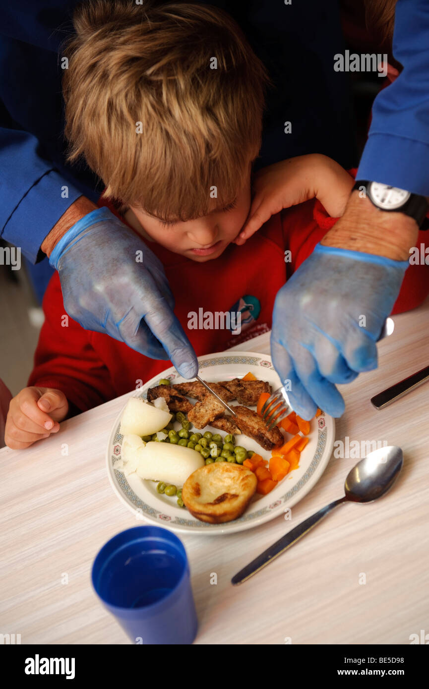 Adulto indossando guanti in lattice blu taglio degli alimenti fino ad un giovane bambino durante la cena di scuola in una scuola primaria, Wales UK Foto Stock