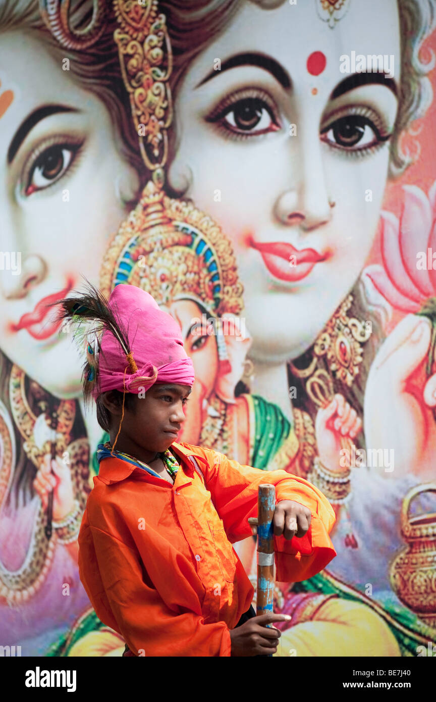 Mendicante indiano ragazzo con lo strumento musicale contro un poster di divinità Indù. Andhra Pradesh, India Foto Stock