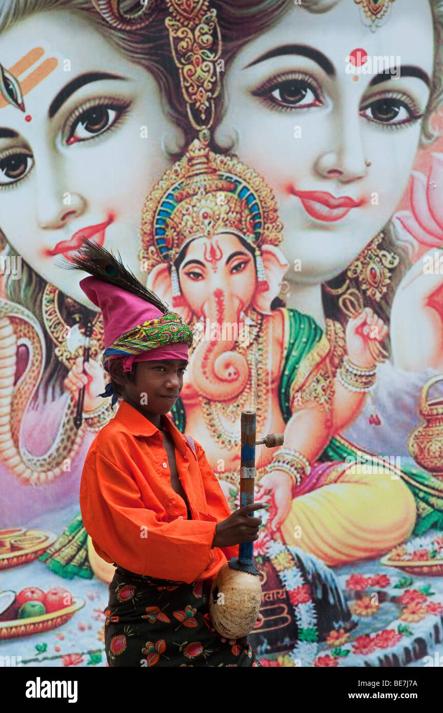 Mendicante indiano ragazzo con lo strumento musicale contro un poster di divinità Indù. Andhra Pradesh, India Foto Stock
