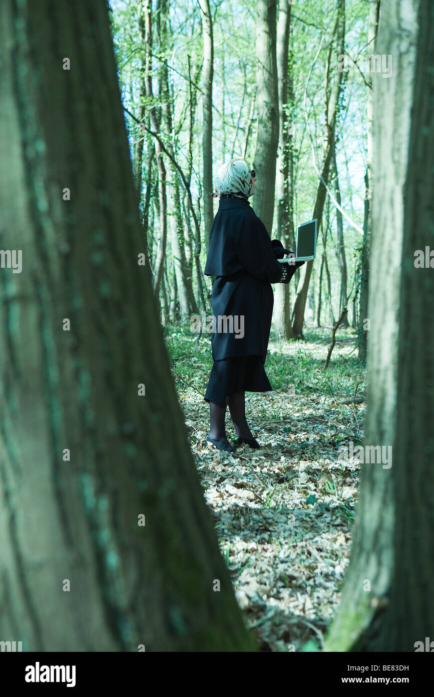 Donna che indossa cappotto e sciarpa di testa in piedi nella foresta, utilizzando il computer portatile Foto Stock