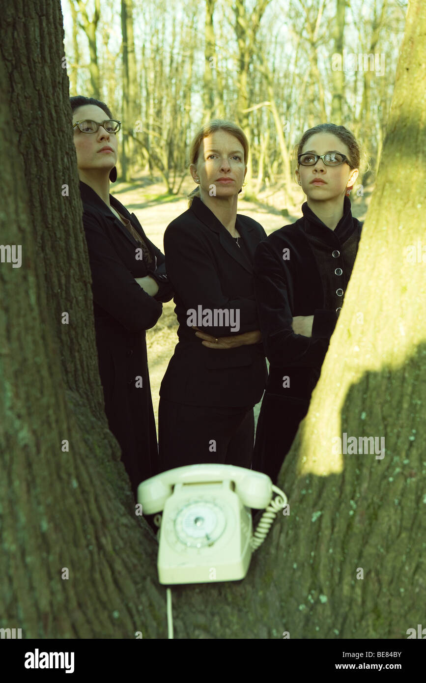 Tre donne in piedi dal tronco di albero con le braccia incrociate, telefono fisso posto in truffatore di tronco Foto Stock