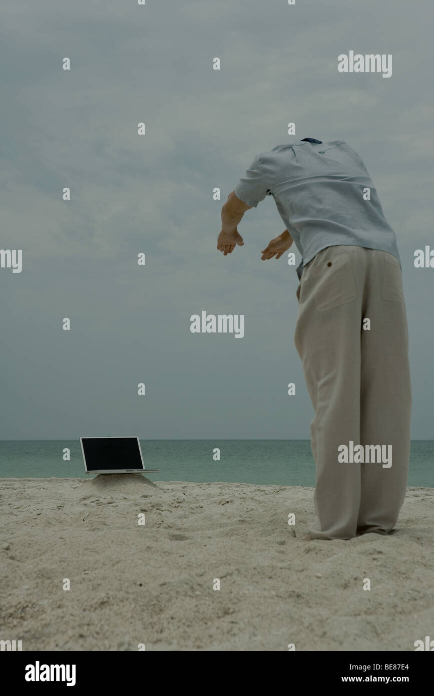 Uomo in piedi sulla spiaggia, piegarsi verso il computer portatile, vista posteriore Foto Stock
