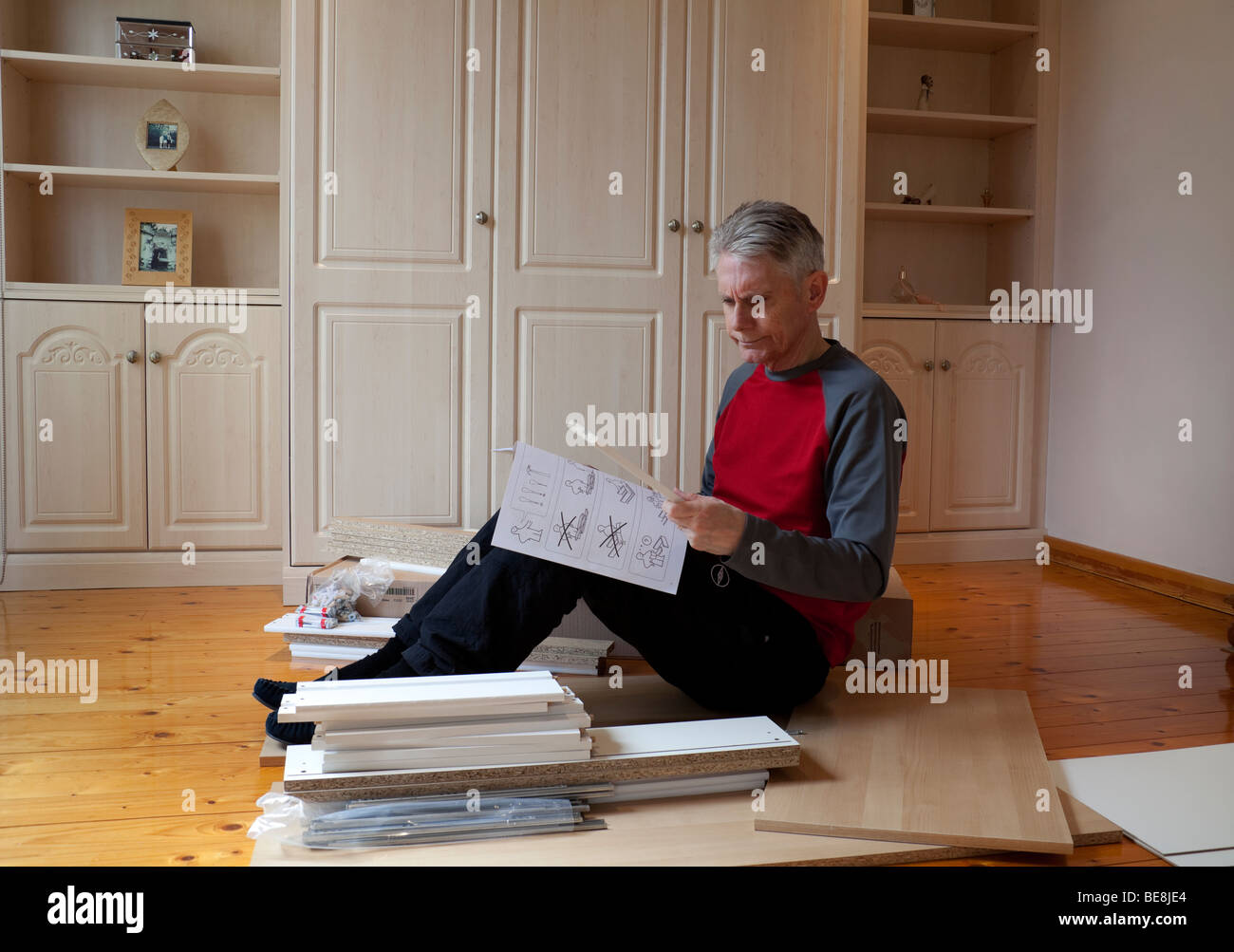 Uomo anziano con i capelli grigi seduta guardando perplesso leggendo foglietto di istruzioni circondato da flat pack mobili FAI DA TE Foto Stock
