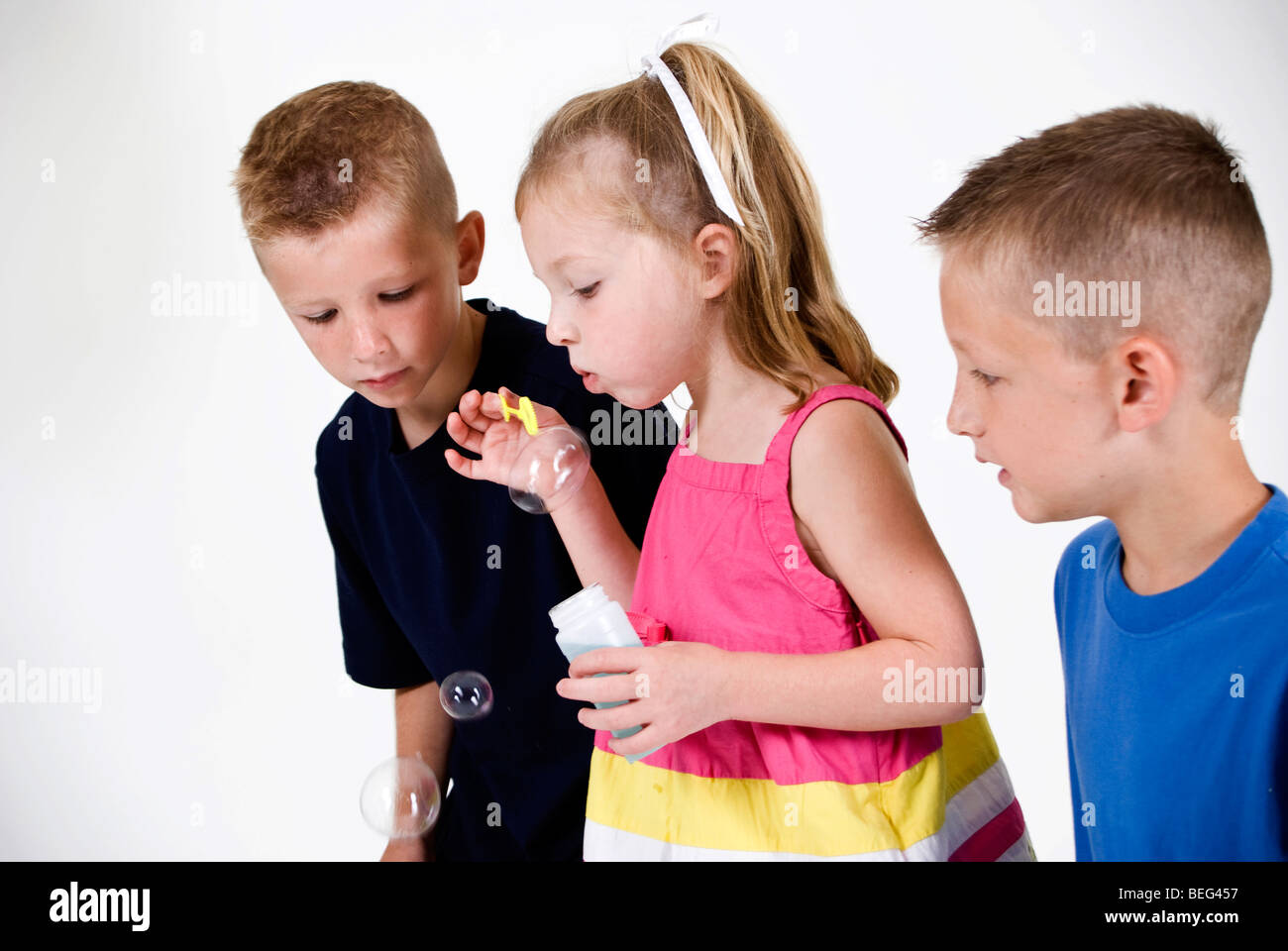 Due fratelli guarda attentamente come loro sorella più piccola crea bolle soffiando acqua saponata attraverso una bacchetta. Foto Stock