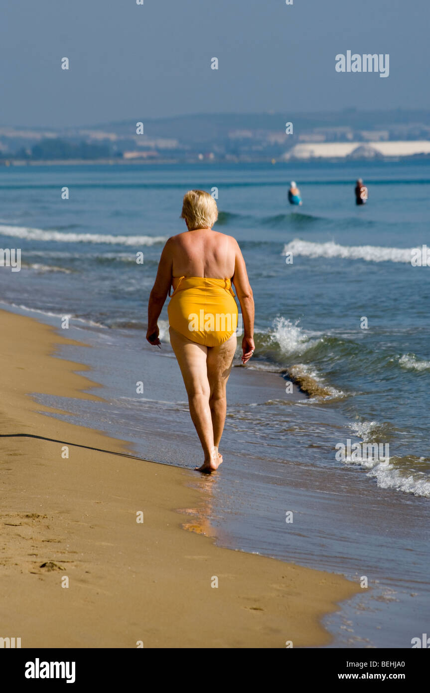 Il sovrappeso donna in un giallo un pezzo unico costume da bagno passeggiando lungo la spiaggia vista da dietro Foto Stock