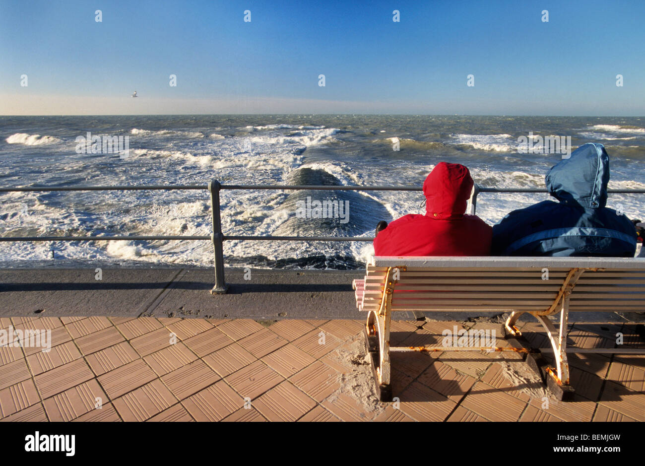 Coppia di turisti su banco sul terrapieno sul mare guardando di onde che si infrangono sulla scogliera in inverno lungo la costa del Mare del Nord Foto Stock