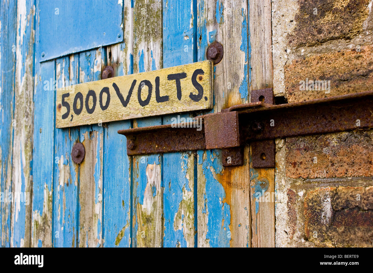 Vecchia porta in legno intemprata con una serratura in metallo arrugginito e un cartello di avvertimento che indica “5.000 VOLT”, che indica l’alta tensione e il pericolo elettrico. Foto Stock