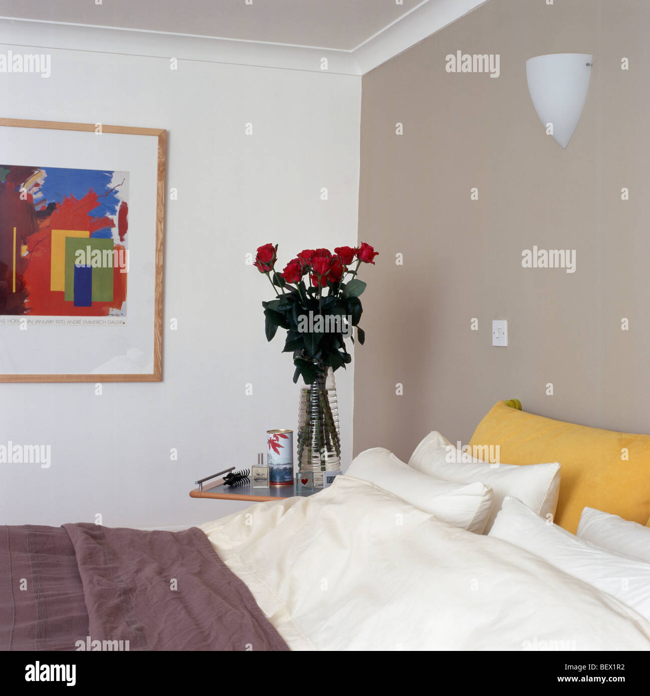 Muro Beige dietro il letto con giallo crema e cuscini in piccole camere da letto moderne con vaso di rose rosse sul comodino Foto Stock