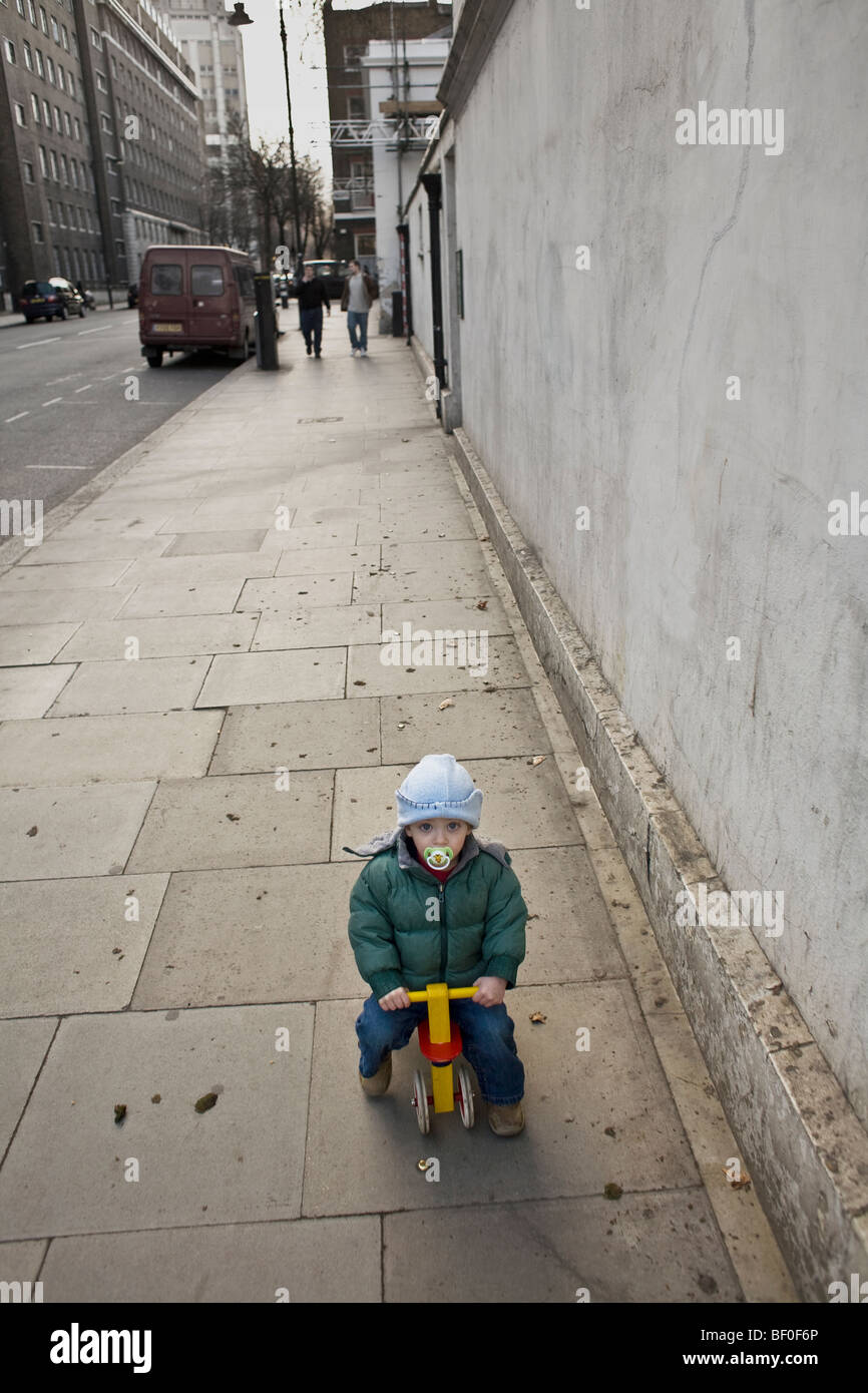 Un giovane bambino con fantoccio su rosso e giallo di triciclo. Marciapiede (marciapiede) tratti dietro. Bloomsbury, Londra, Regno Unito. Foto Stock