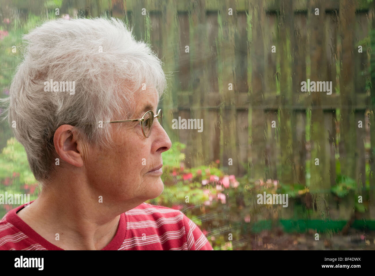Senior donna anziana con un espressione indisponente guardando fuori attraverso una finestra da sola e la sensazione di essere premuta da un giorno di pioggia. Regno Unito depressione salute mentale Foto Stock