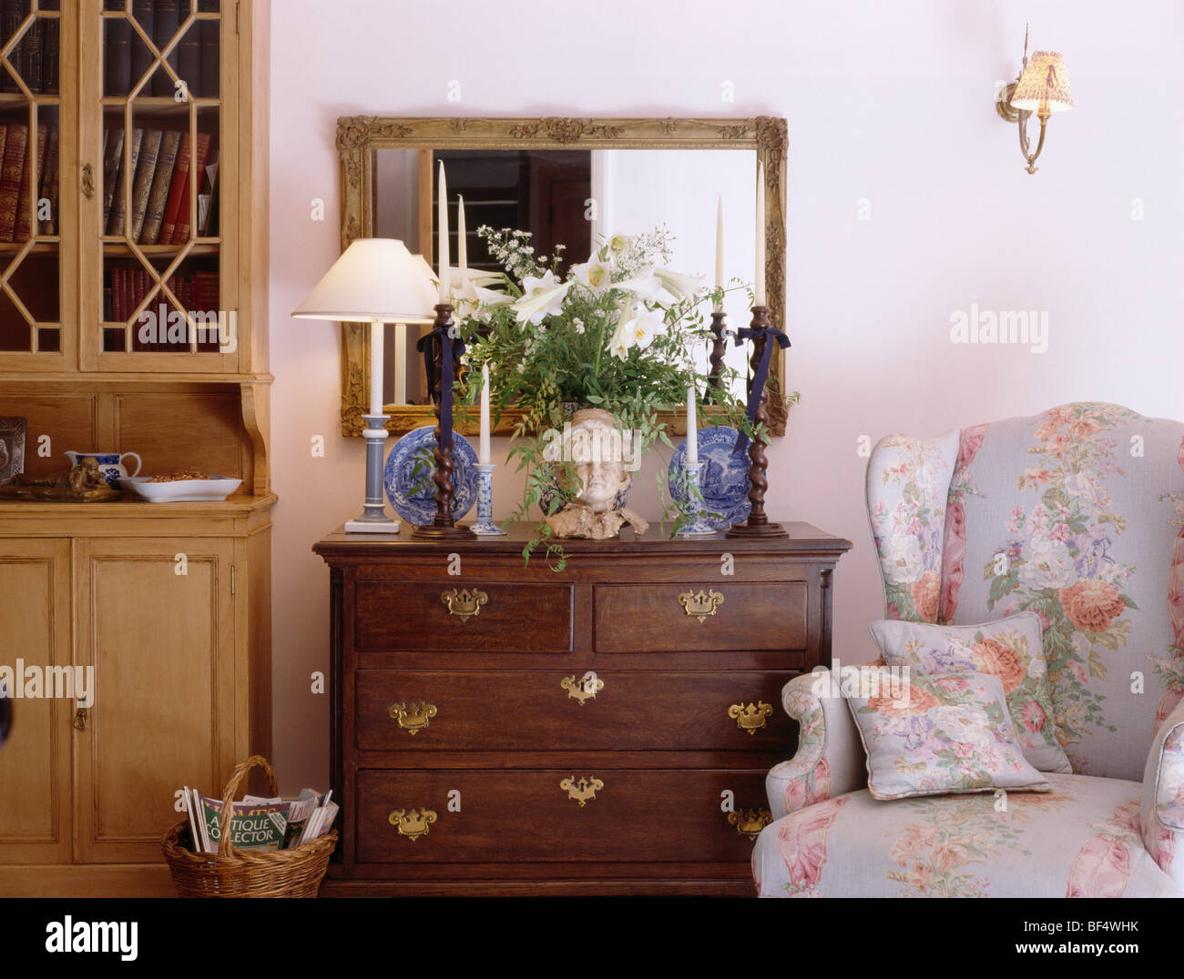 Specchio sopra lampada accesa e vaso di fiori sul petto di antiquariato-di-cassetti accanto ala floreale-sedia in cottage soggiorno Foto Stock