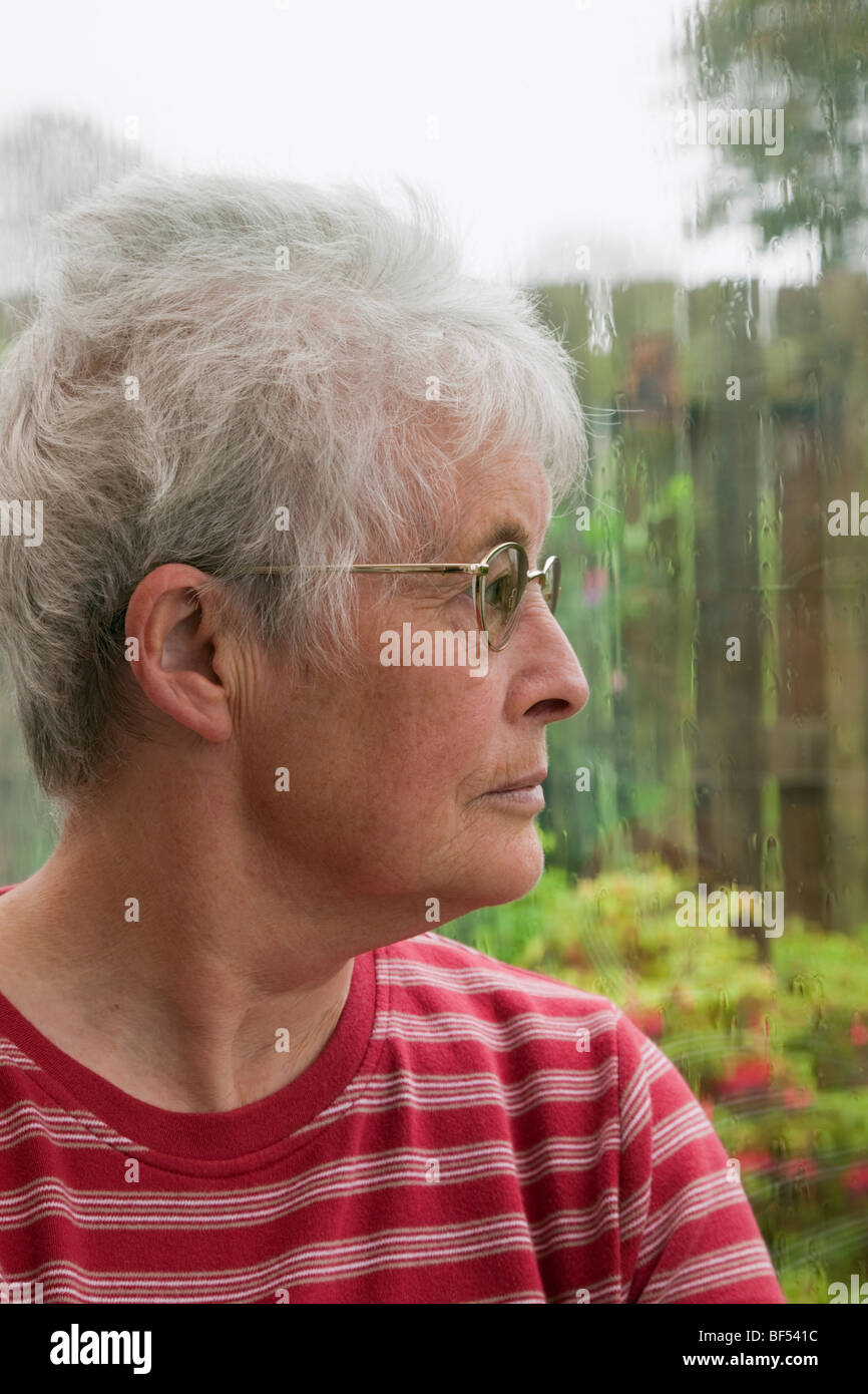 Senior donna con una espressione indisponente guardando fuori attraverso una finestra in un giorno di pioggia. Regno Unito Gran Bretagna Foto Stock