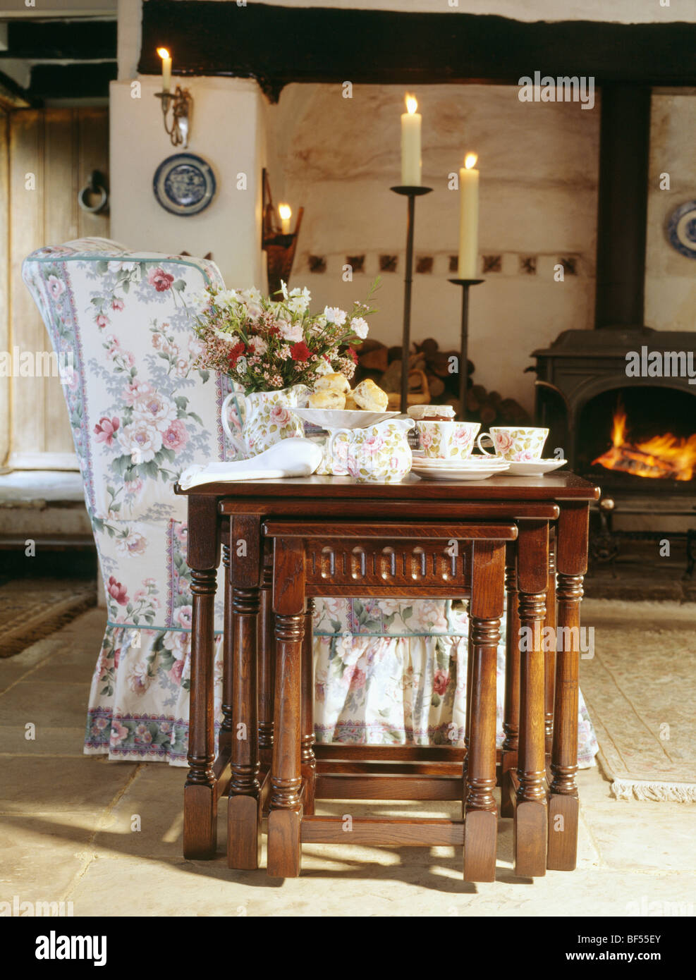 Oak nest-di-tabelle accanto al divano floreale in cottage soggiorno con le candele accese in alti candelabri accanto alla stufa nero Foto Stock