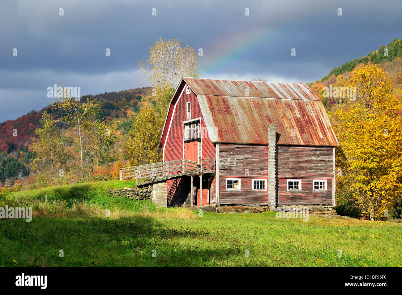 Un vecchio fienile nel Vermont con fogliame autunnale che lo circonda. C'è anche un cielo tempestoso con un arcobaleno sopra il fienile. Vicino a Hancock, Vermont. Foto Stock