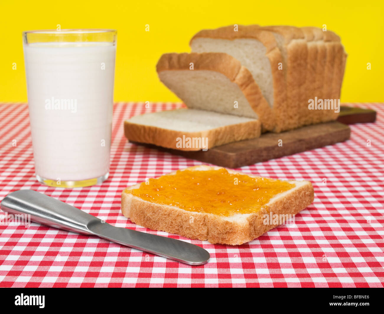 Una fetta di pane spalmato con marmellata accanto a un filone di pane e un bicchiere di latte. Foto Stock