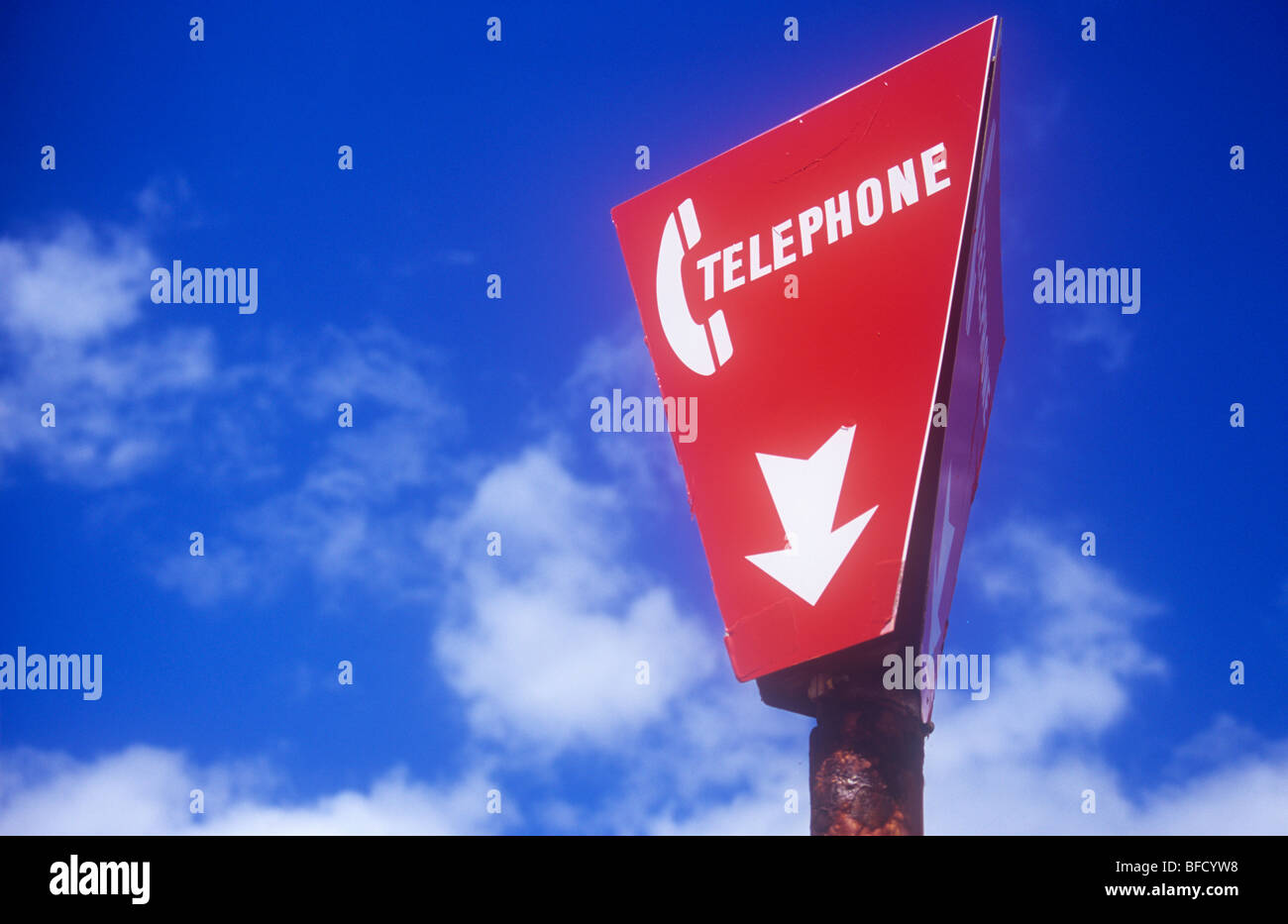 Segno rosso con icona del telefono e il telefono di parola con la freccia rivolta verso il basso appollaiato sul palo arrugginito sotto il cielo blu con nuvole bianche Foto Stock