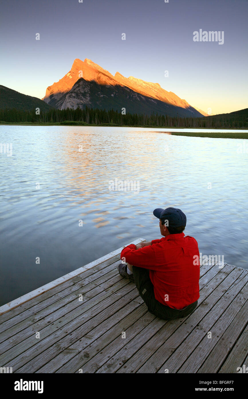 Maschio adulto seduto sul dock Vermillion lago al tramonto con Mount Rundle in background, il Parco Nazionale di Banff, Alberta, Canada. Foto Stock